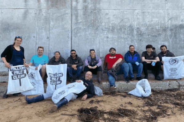 Group Of People Sitting By A Sea Wall Holding “million Mile Clean” Bags After A Beach Clean Up.